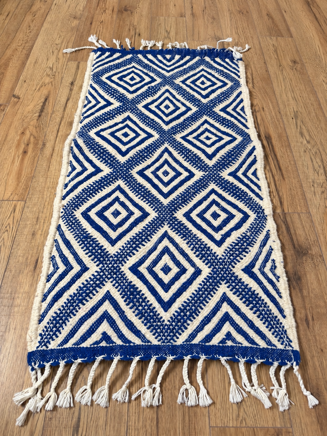 Petit tapis en laine écru et bleu à motifs losanges et franges nouées, présenté à plat sur un parquet
