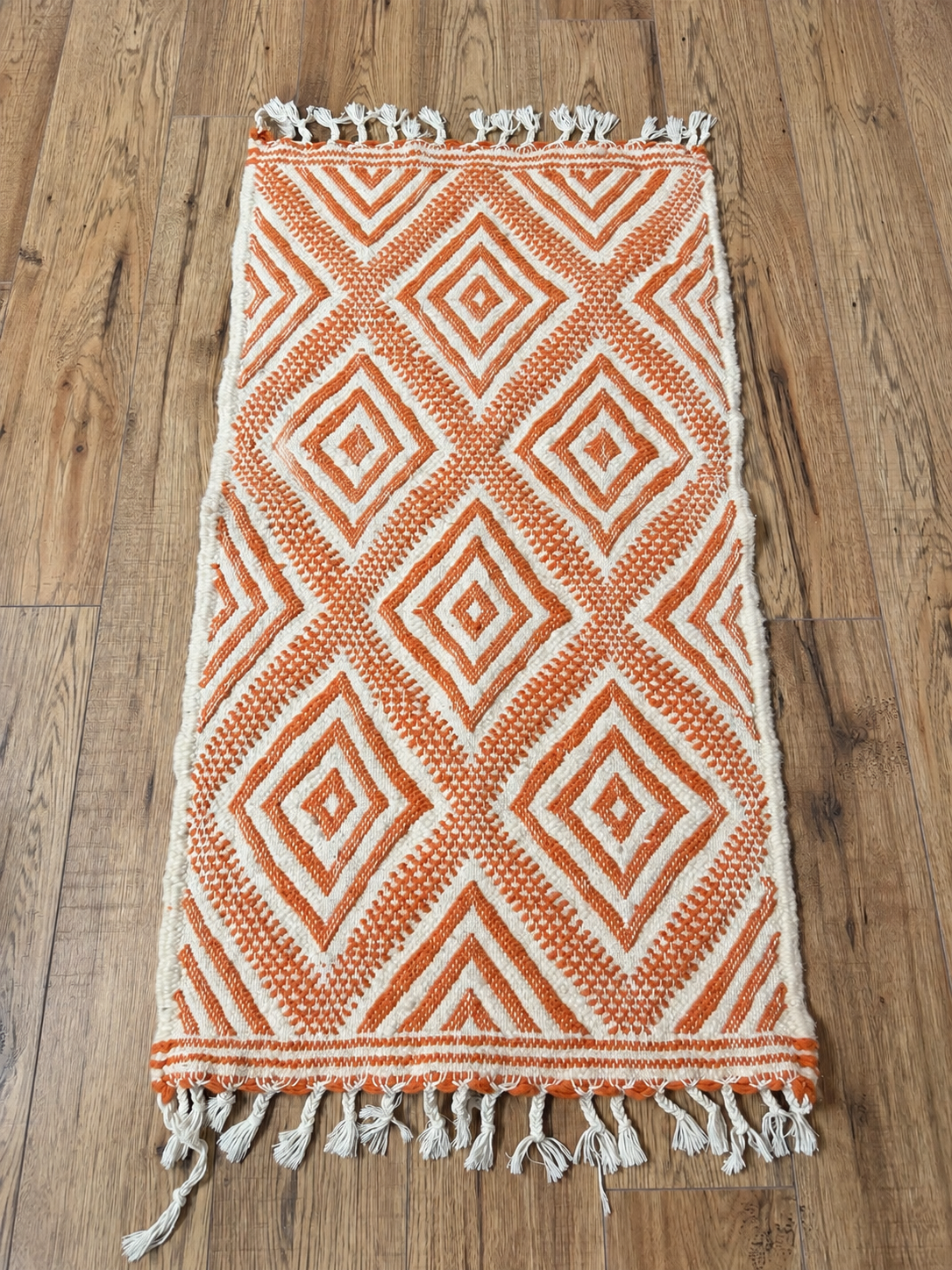 Petit tapis en laine écru et orange à motifs losanges et franges nouées, présenté à plat sur un parquet