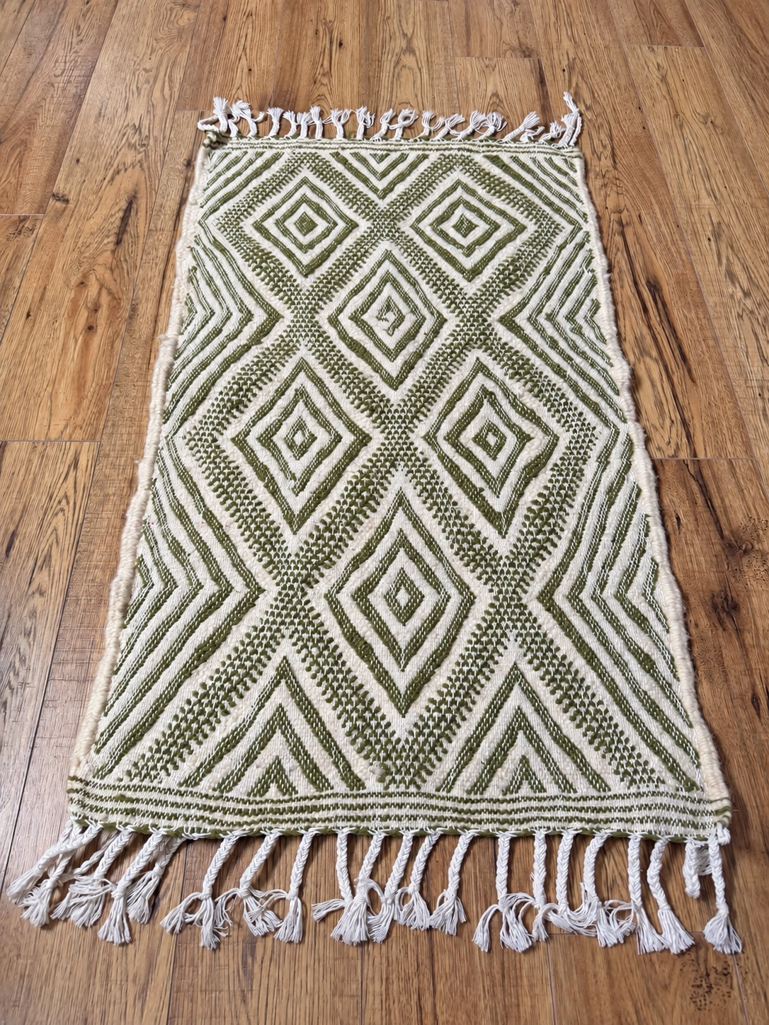Petit tapis en laine écru et vert olive à motifs losanges et franges nouées, présenté à plat sur un parquet